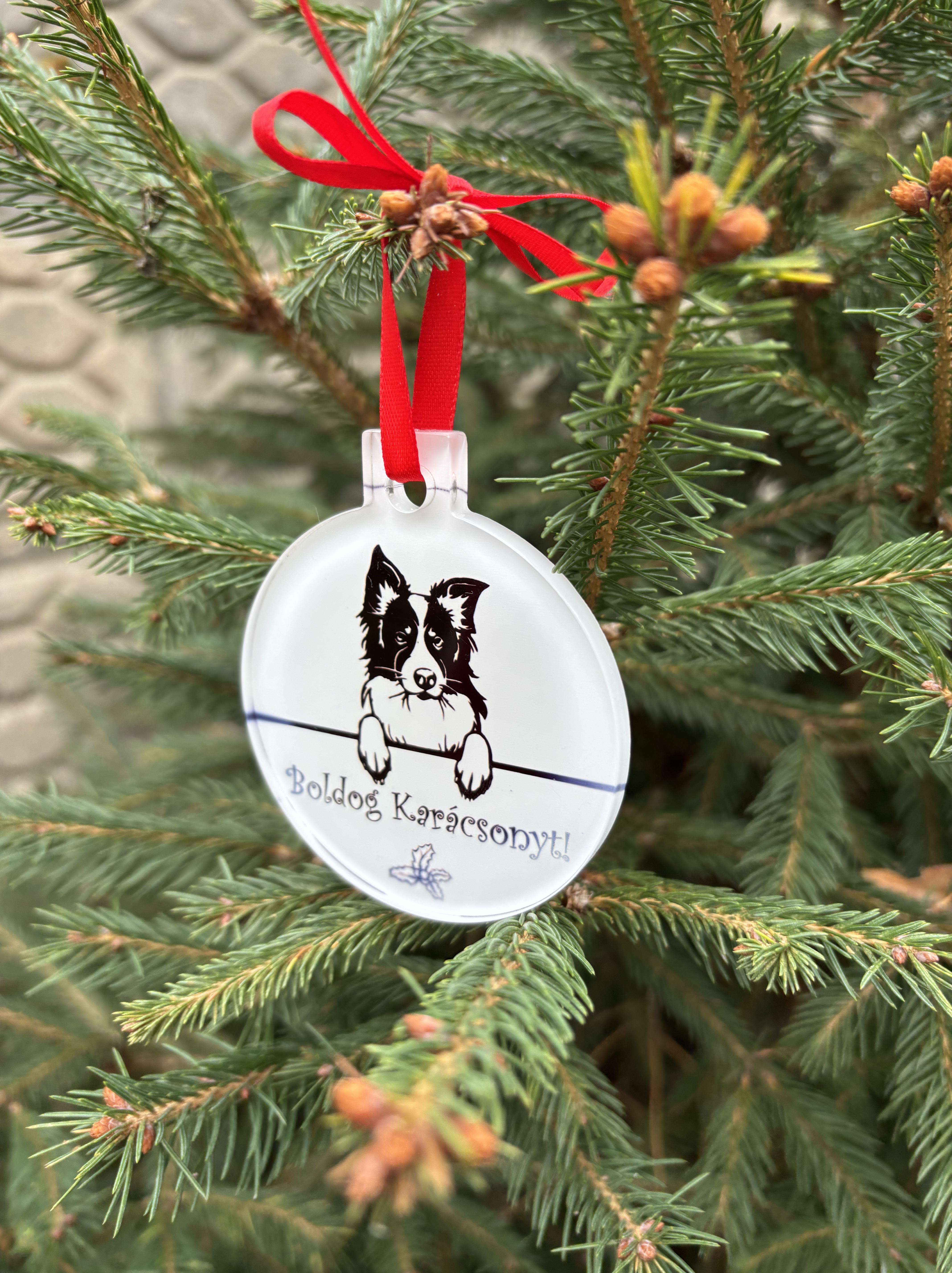 Akril karácsonyfa dísz kutyával, Boldog karácsonyt felirattal, fenyőfára akasztva, piros masnival megkötve.